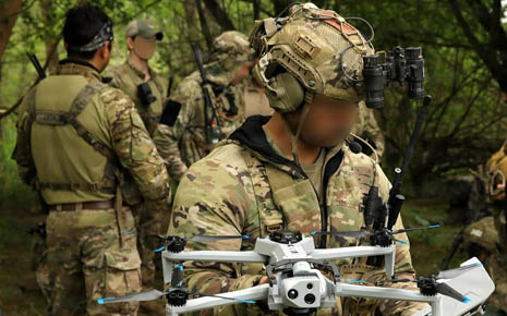 People wearing camouflage military gear stand in a wooded area with one person holding a drone and wearing a helmet with night vision goggles. Description generated by AI