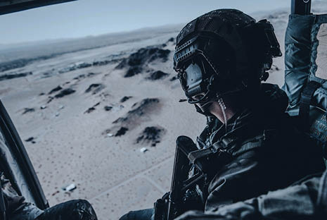 A person wearing tactical gear and a helmet is sitting inside a helicopter looking out over a desert landscape. Description generated by AI