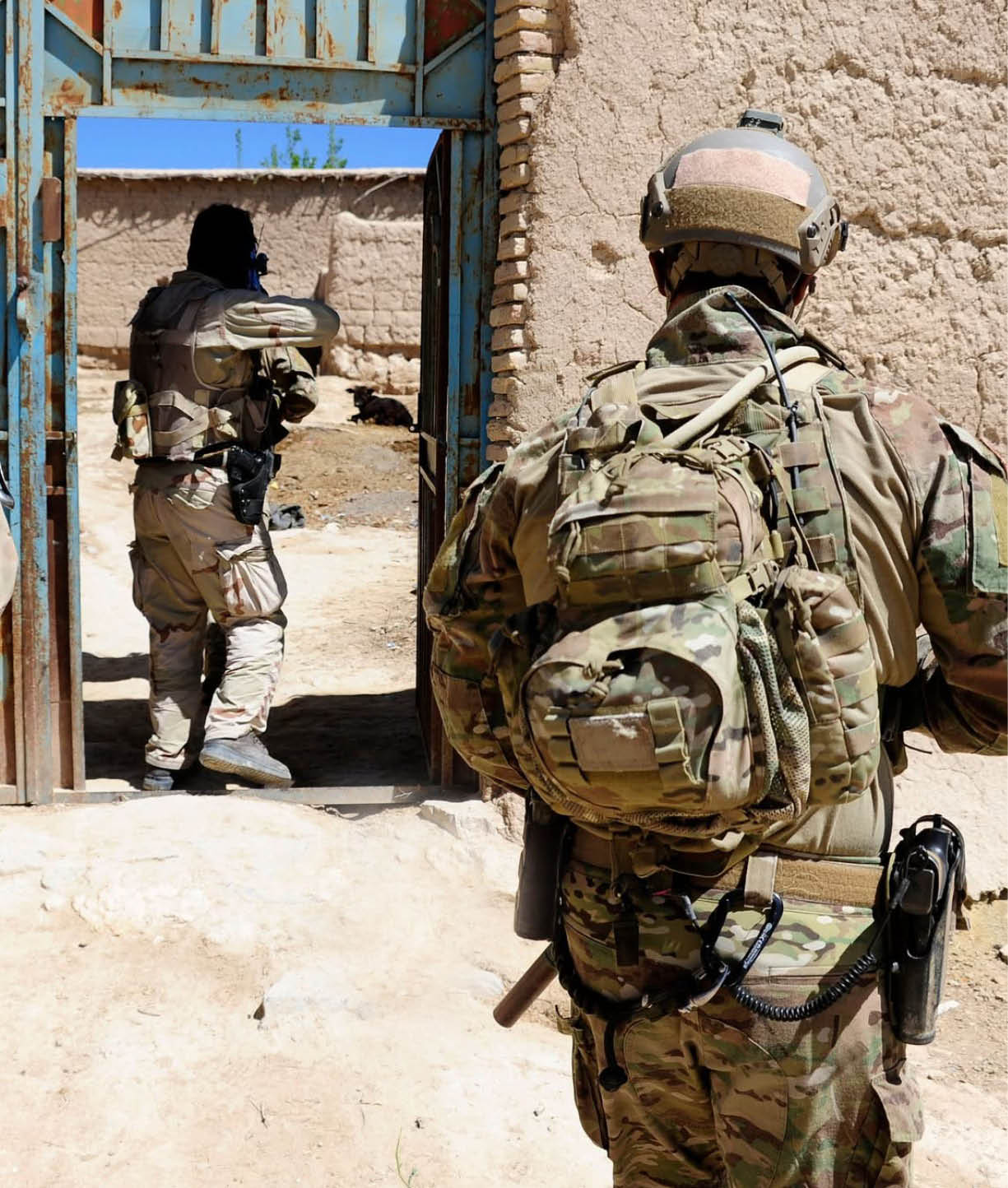 Two people in military camouflage gear and helmets are positioned near a doorway in a sandy, outdoor environment. Description generated by AI