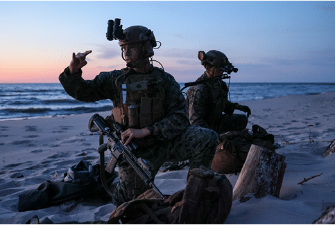 Two men in military uniforms are sitting on the beach, one pointing at the other. They have backpacks and a handbag nearby. AI generated content