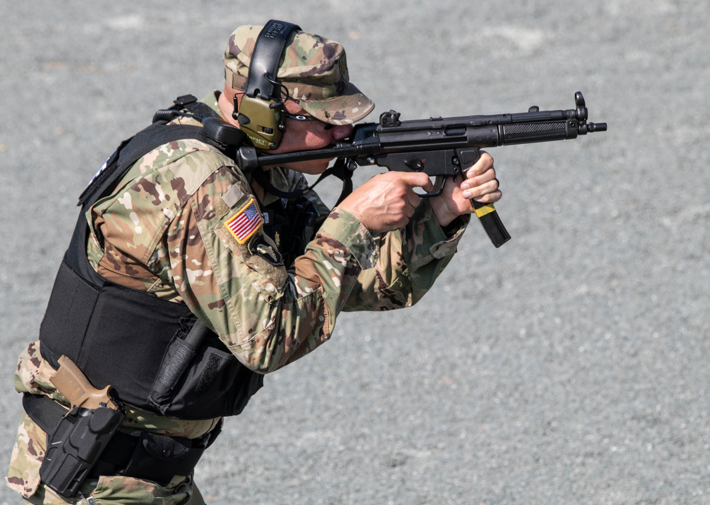 A person wearing camouflage clothing and protective headphones is aiming a black rifle while crouching on a gravel surface. Description generated by AI