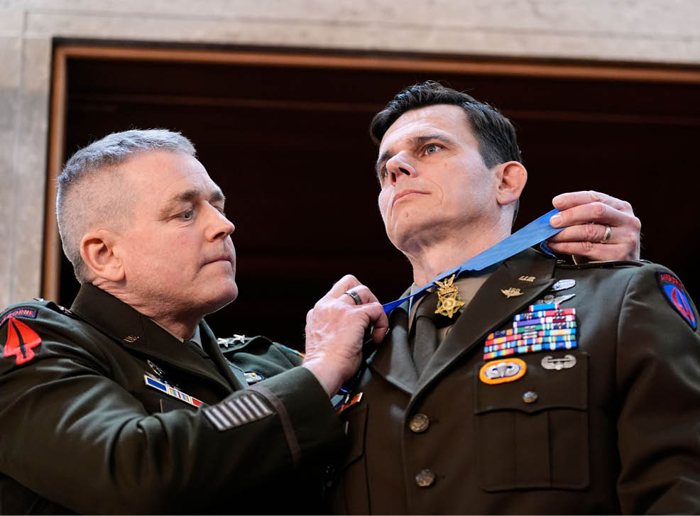 An adult in a military uniform is placing a medal around the neck of another adult in a military uniform inside a building. Description generated by AI