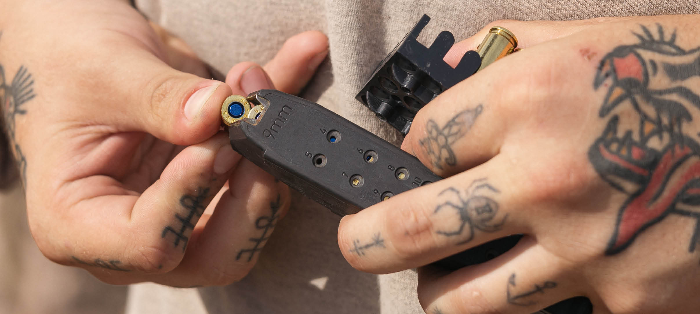 Person with tattoos on their hands loading a bullet into a black magazine against a neutral background. Description generated by AI