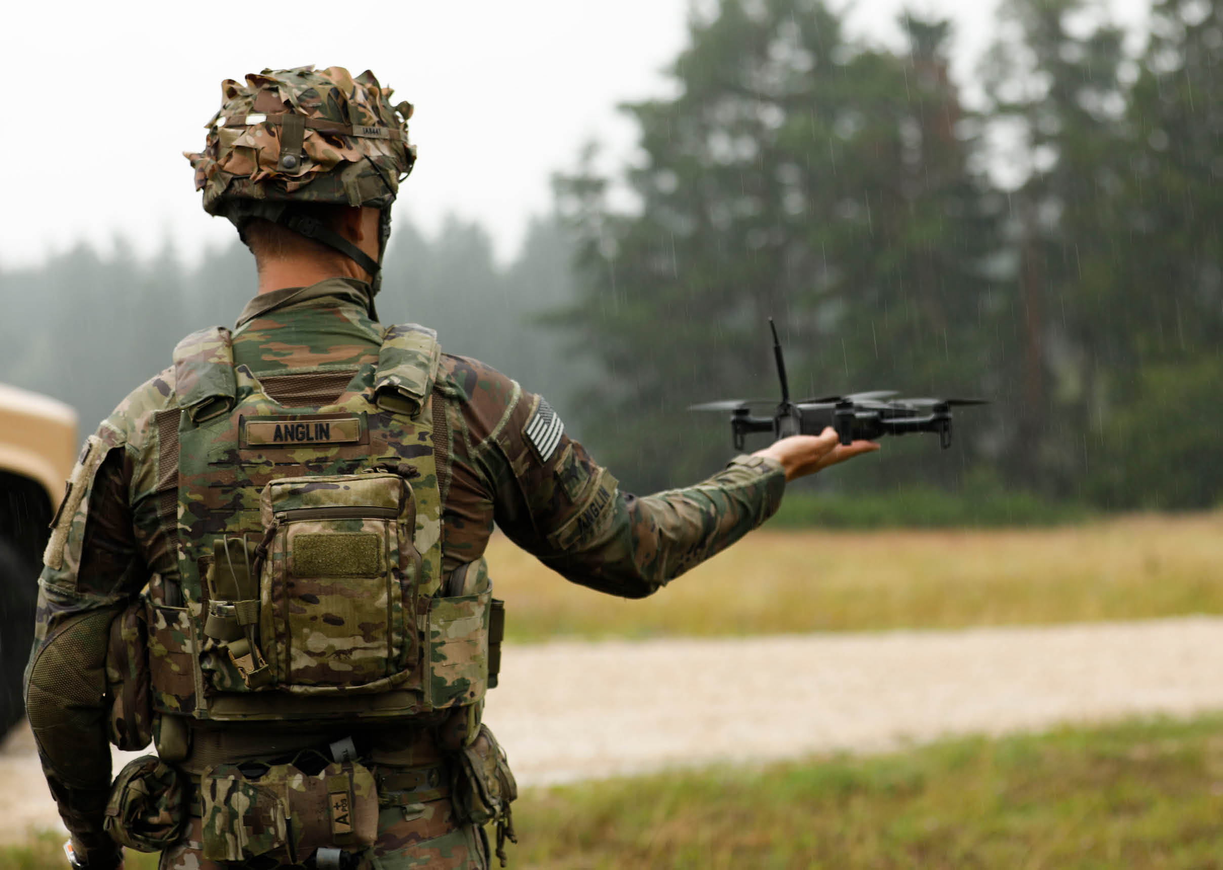 A person in military camouflage uniform and helmet is holding a small drone in an outdoor setting with trees and grass in the background. Description generated by AI