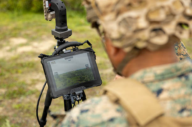 A person wearing camouflage clothing and a helmet is looking at a mounted screen displaying an outdoor landscape. Description generated by AI