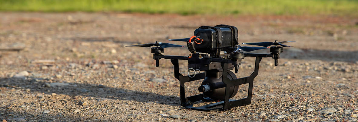 A black drone with four propellers is resting on a gravel surface with grass and trees in the background. Description generated by AI