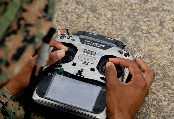 A person wearing camouflage clothing is holding and operating a remote control device with multiple buttons and joysticks. Description generated by AI