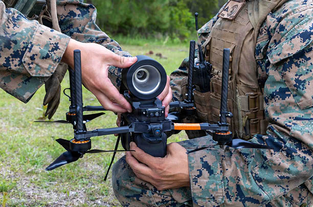 Two people wearing camouflage uniforms are holding and inspecting a black drone with four propellers outdoors on grass. Description generated by AI