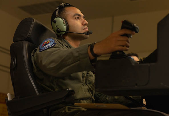 An adult wearing a headset and a green flight suit is seated in a chair operating a joystick in a dimly lit room. Description generated by AI