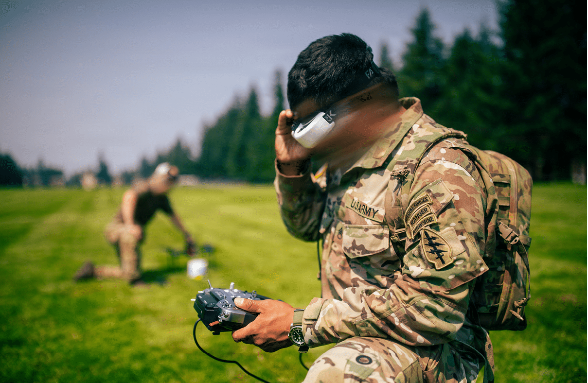 A person in military camouflage uniform is holding a radio near their mouth while another person in similar attire is blurred in the background on a grassy field. Description generated by AI
