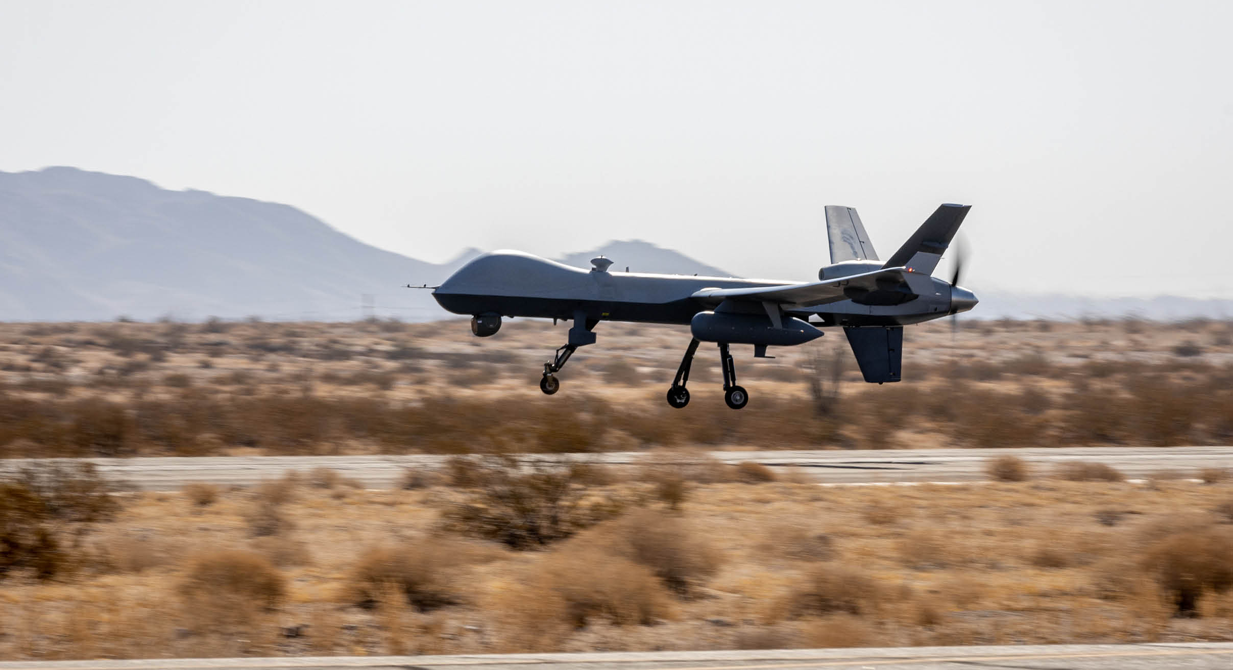 A military drone is flying low over a desert landscape with mountains in the background. Description generated by AI
