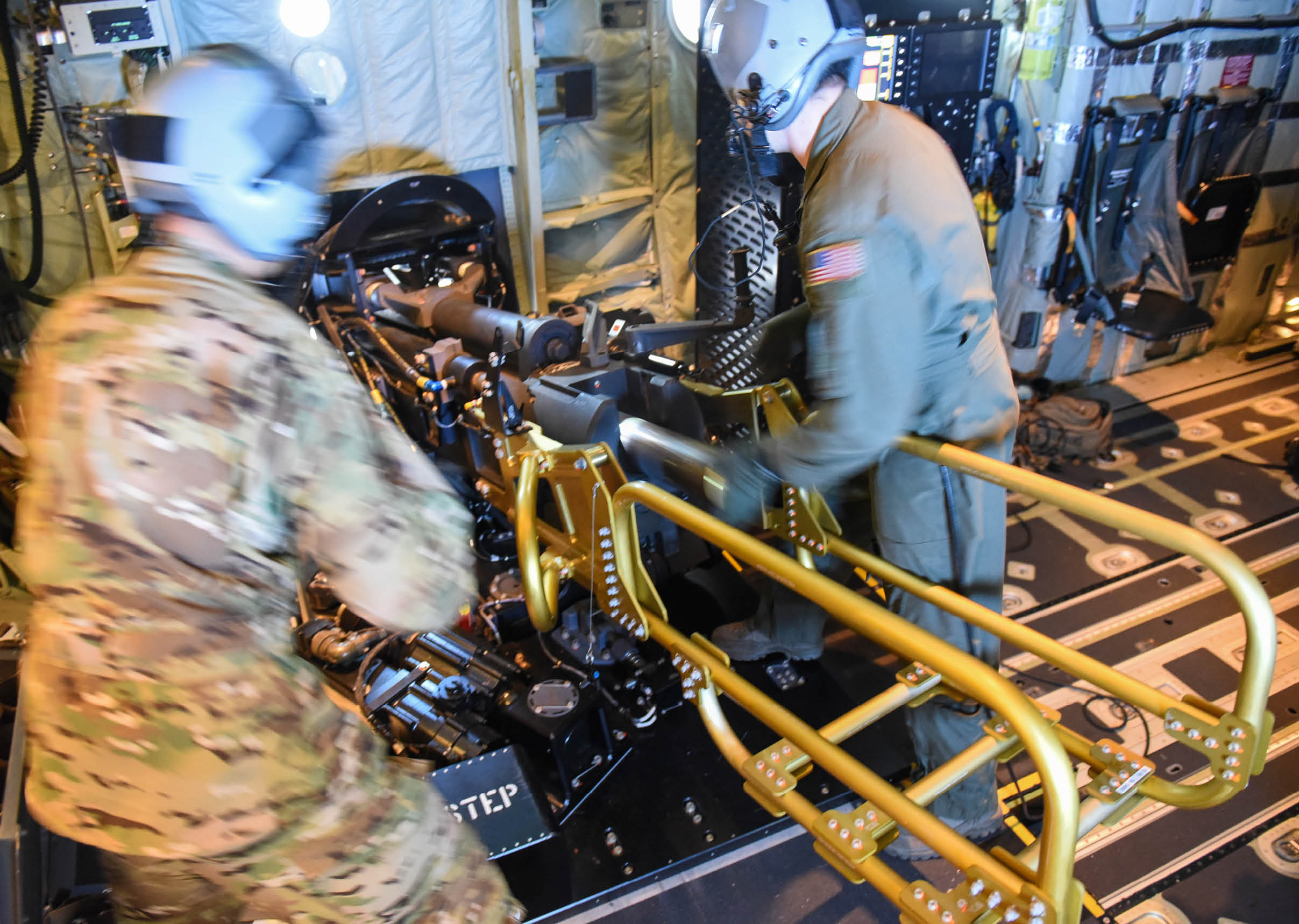 Two people wearing helmets and military-style clothing operate a mounted machine gun inside a vehicle or aircraft. Description generated by AI