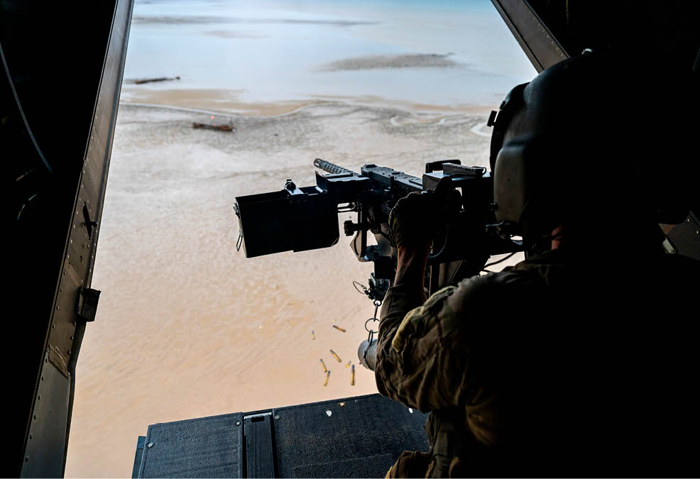 A person wearing a helmet is operating a mounted machine gun from the open rear of a helicopter flying over a sandy or muddy terrain. Description generated by AI