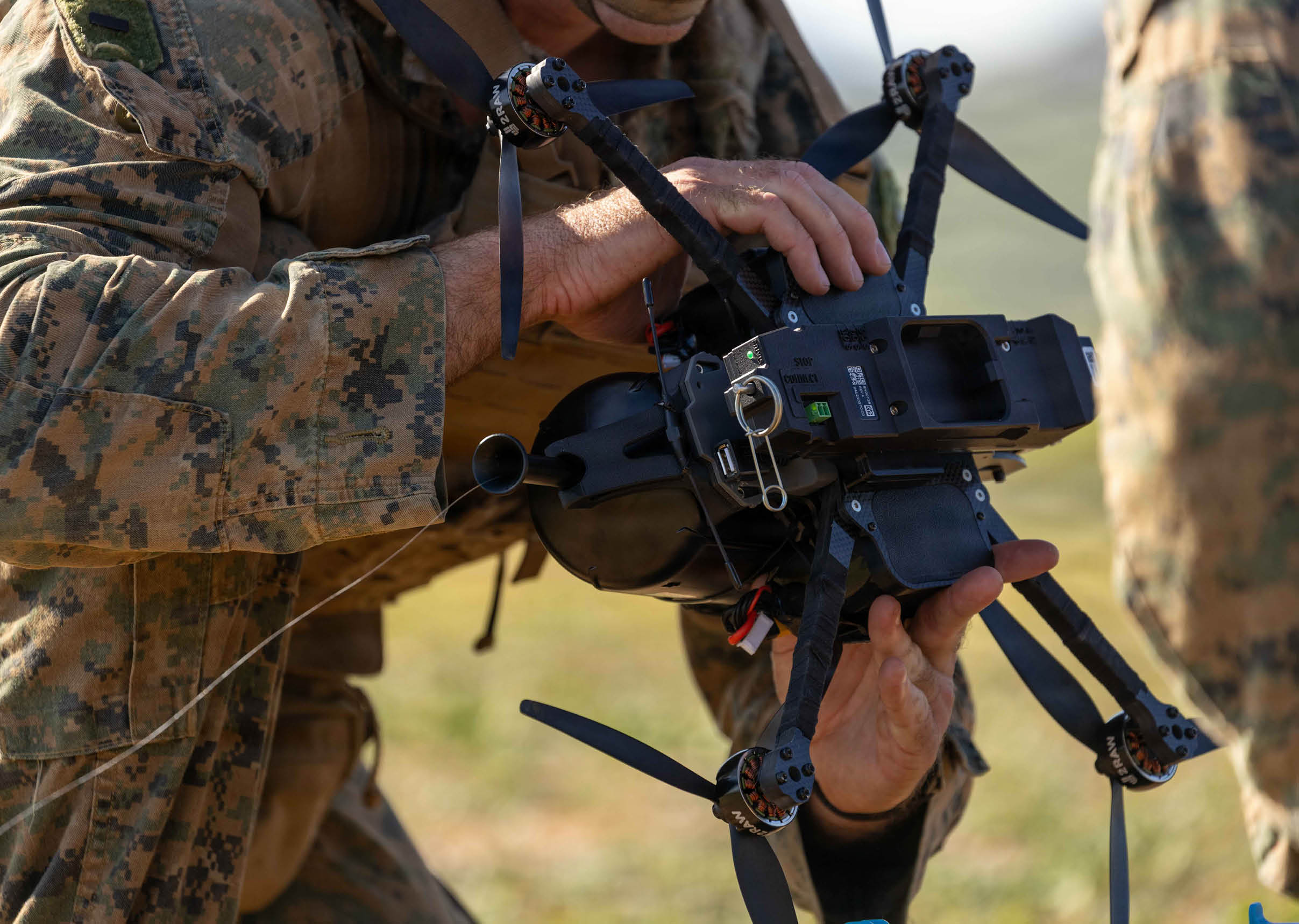 A person wearing camouflage clothing is holding and inspecting a black drone with four propellers outdoors. Description generated by AI