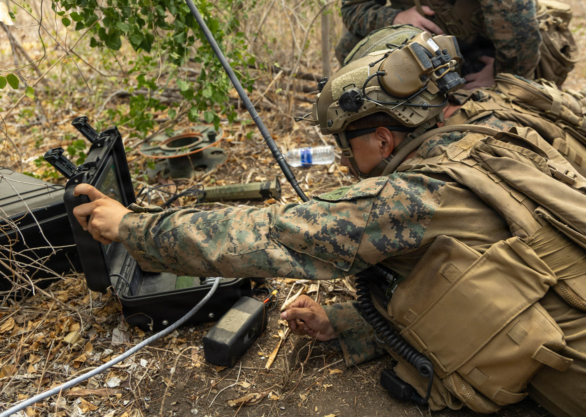 A person in military gear is lying on the ground outdoors, reaching into a black case while handling equipment near some cables and foliage. Description generated by AI