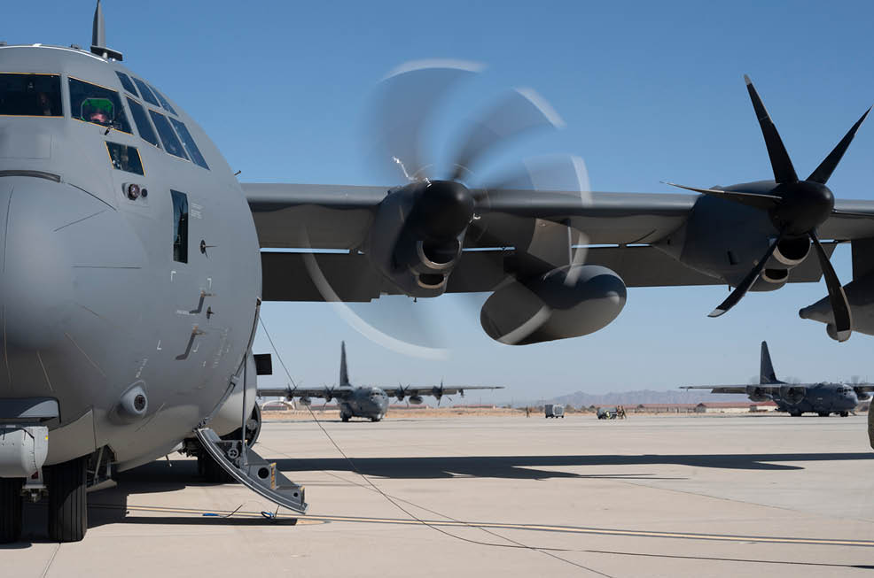 A military transport aircraft with spinning propellers is parked on a runway with other similar aircraft visible in the background under a clear blue sky. Description generated by AI