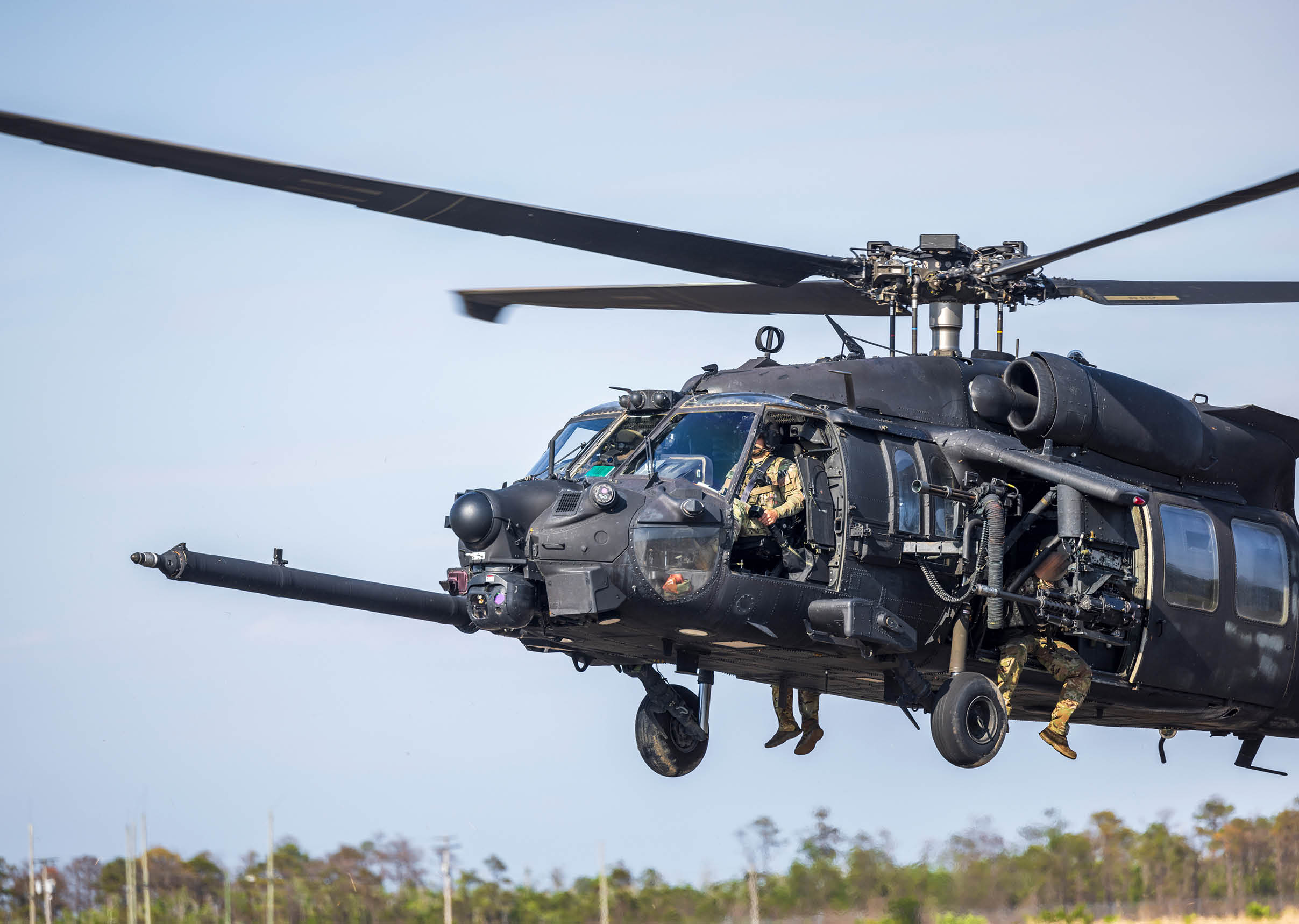 A black military helicopter is flying low over a field with two people visible inside and one person hanging from the open side door. Description generated by AI