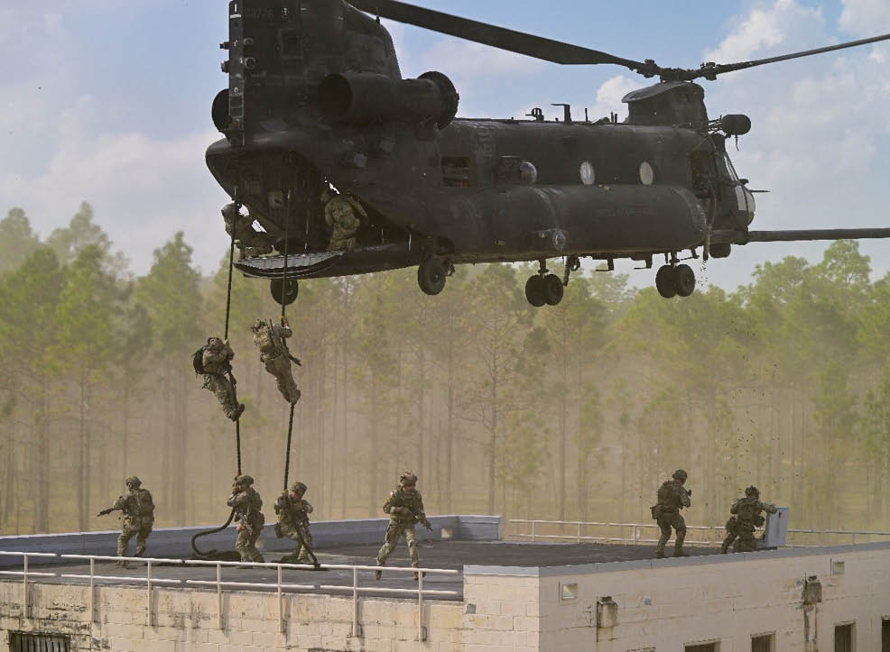 Several people in military gear are rappelling from a helicopter onto a rooftop while others stand on the rooftop in a forested area. Description generated by AI