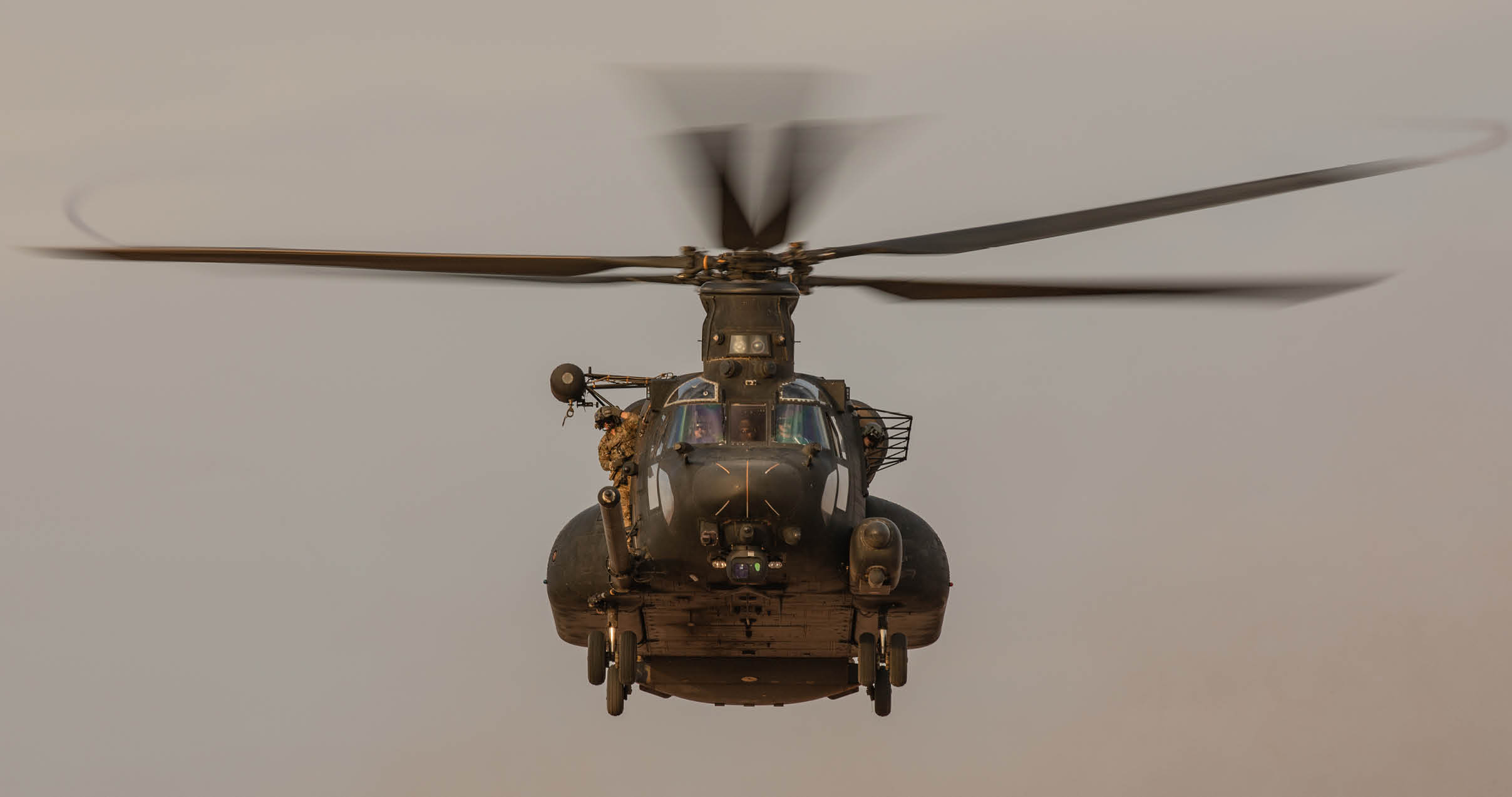 A military helicopter is captured head-on with its rotor blades spinning against a plain sky background. Description generated by AI