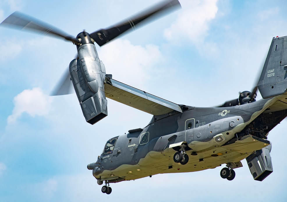 A military tiltrotor aircraft is flying against a partly cloudy sky with its rotors in motion. Description generated by AI