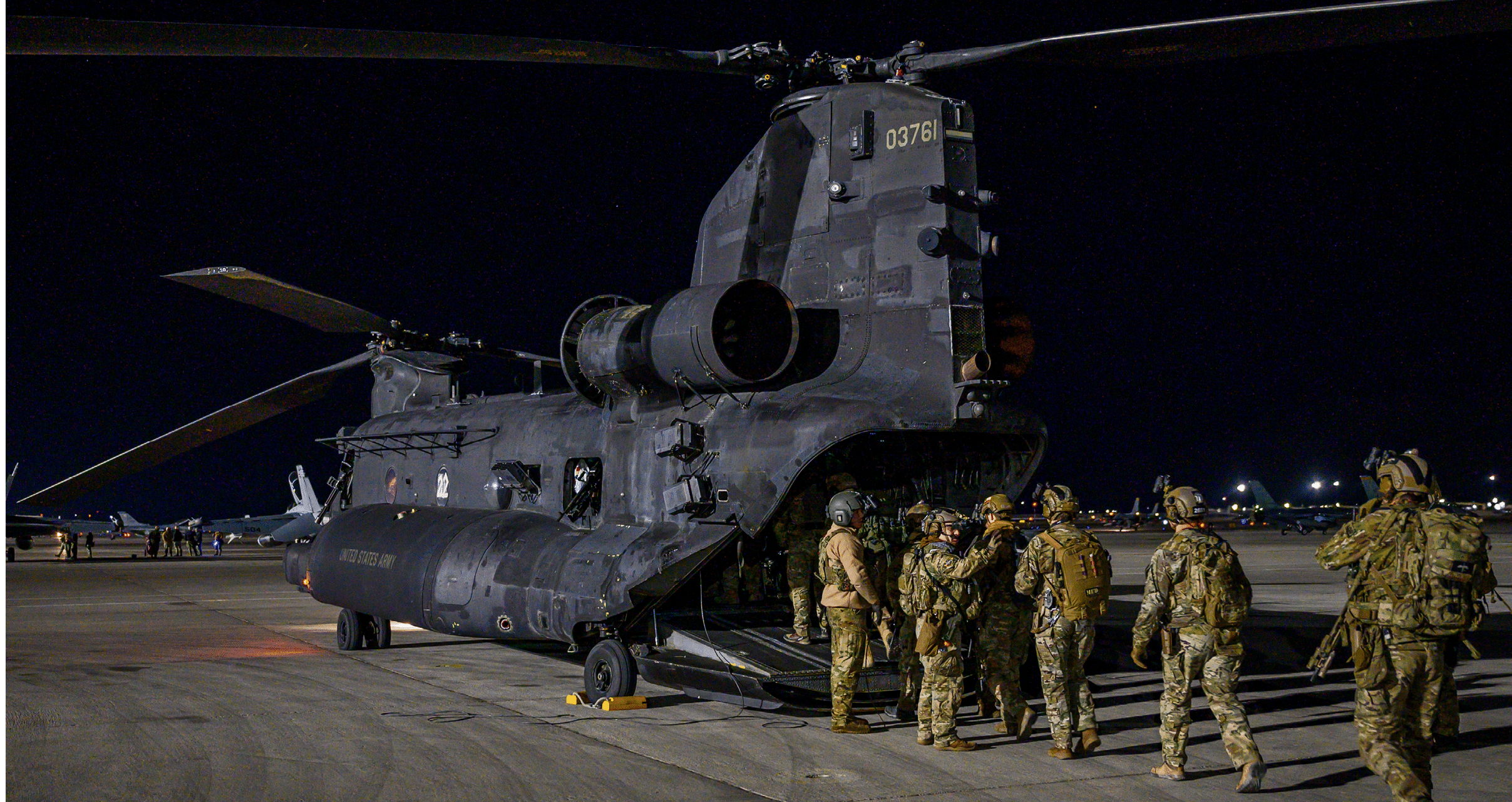 A group of people in military gear are walking towards a large, dark-colored helicopter on an airfield at night. Description generated by AI