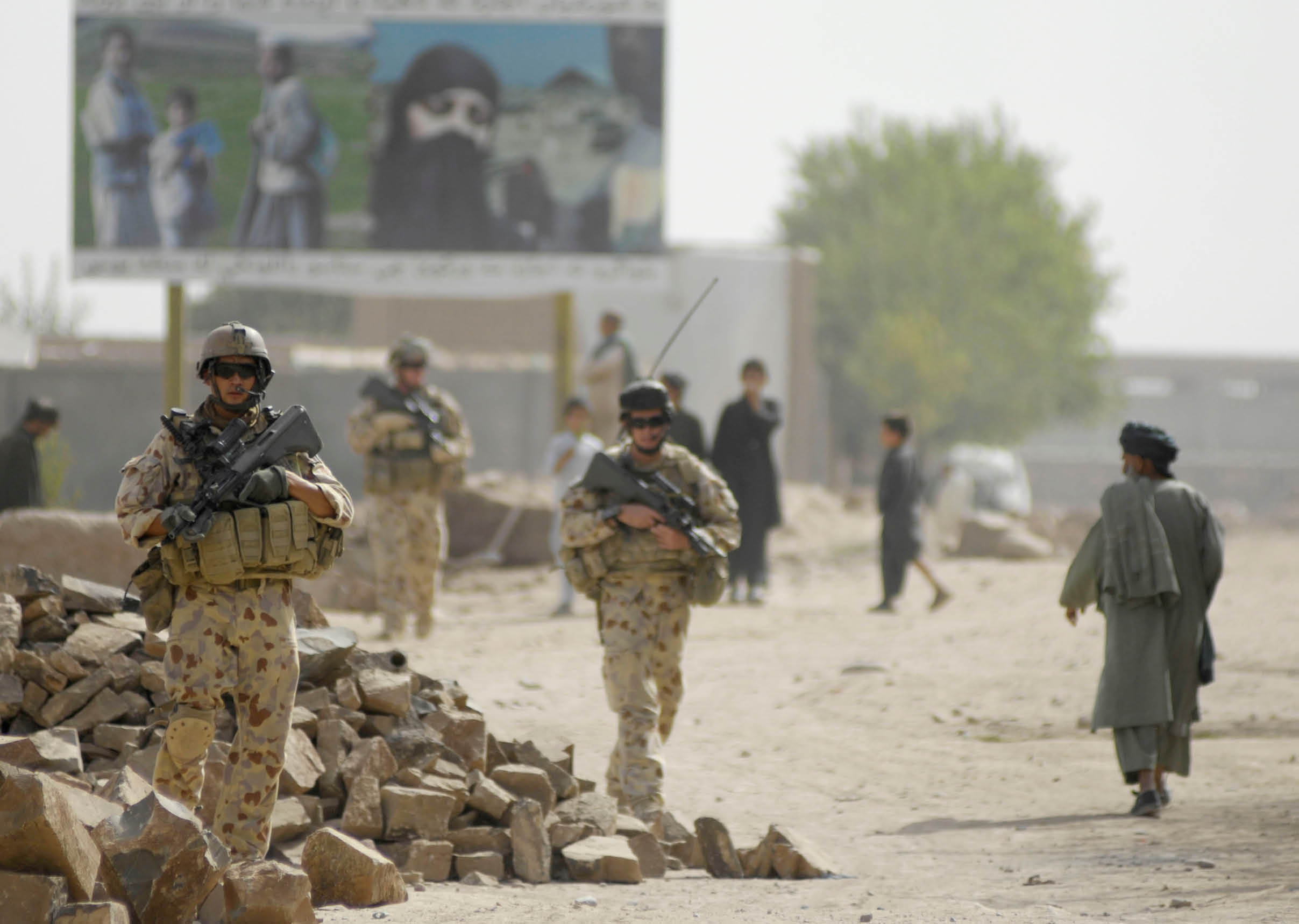 Armed adults in military uniforms walk along a dirt road near a pile of rocks while other people in traditional clothing stand or walk nearby with a large billboard in the background. Description generated by AI