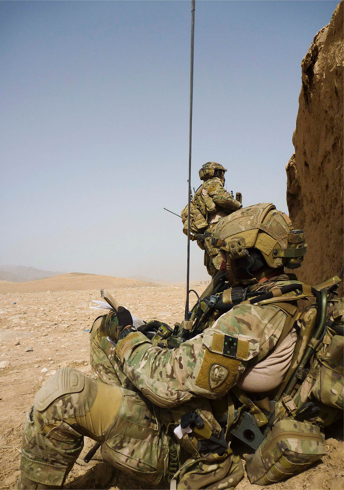 Two people in military gear are in a desert setting, with one person holding a notebook and the other standing nearby. Description generated by AI