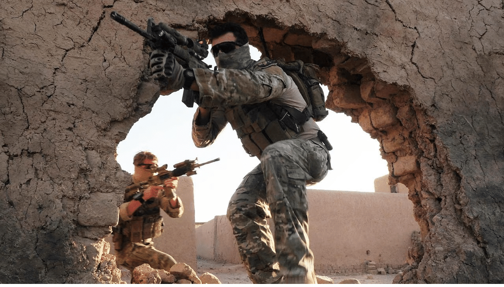 Two people in military camouflage gear are aiming rifles while positioned near a wall in an outdoor setting. Description generated by AI