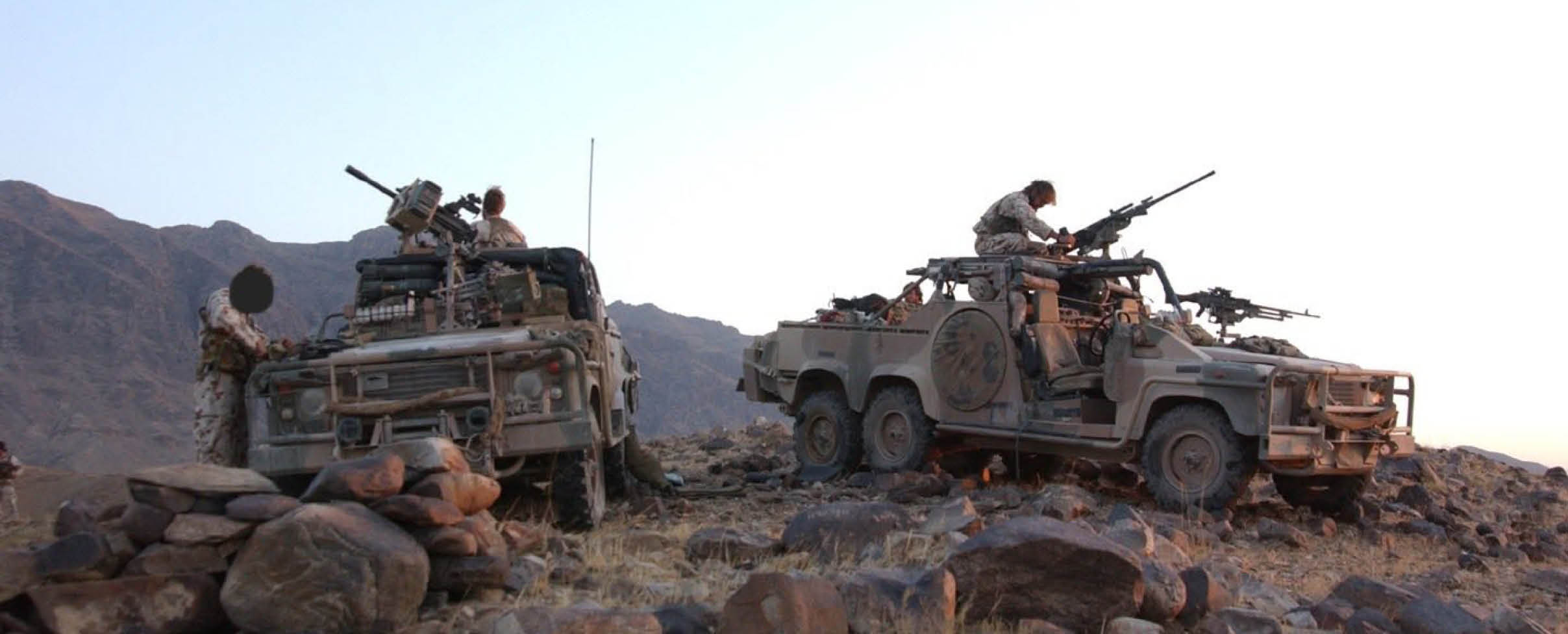 Two military vehicles equipped with mounted guns are positioned on rocky terrain with several people in military uniforms standing or sitting on them against a backdrop of mountains and clear sky. Description generated by AI
