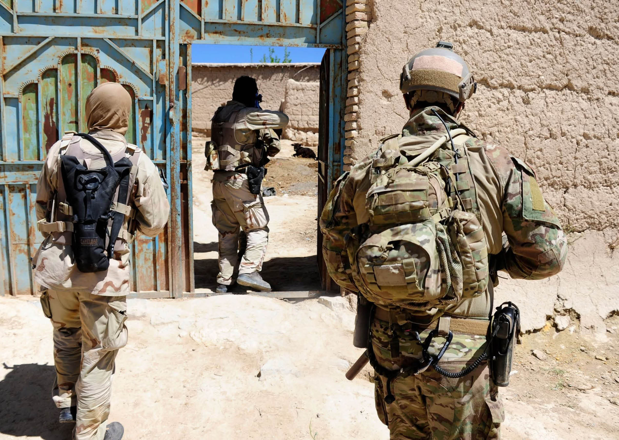 Three people in military gear are walking through a sandy area with a metal gate and mud-brick walls under a clear blue sky. Description generated by AI