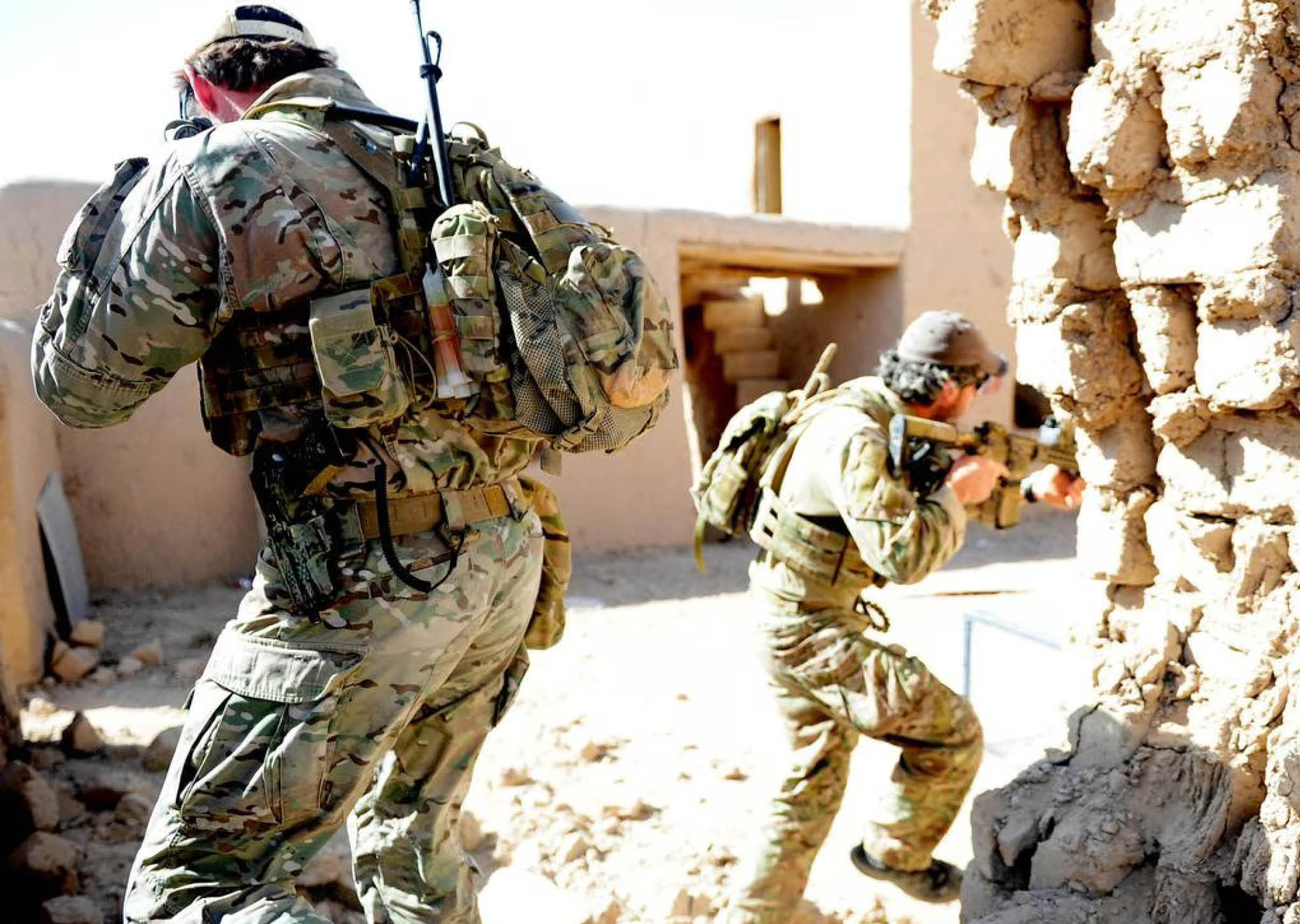 Two people in camouflage military uniforms with backpacks and rifles are positioned near a damaged wall in a sunlit, sandy environment. Description generated by AI