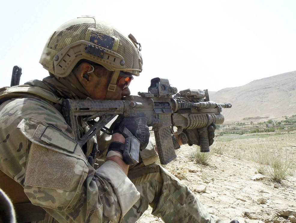 A person wearing tactical gear and a helmet is aiming a rifle equipped with various attachments in a desert-like outdoor setting. Description generated by AI