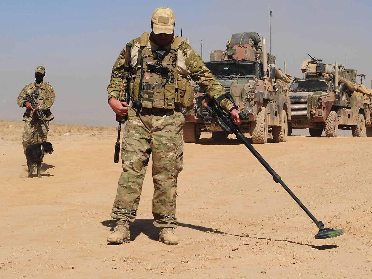 A person in military camouflage gear uses a metal detector on a dusty road while another person with a dog and armored vehicles are in the background. Description generated by AI