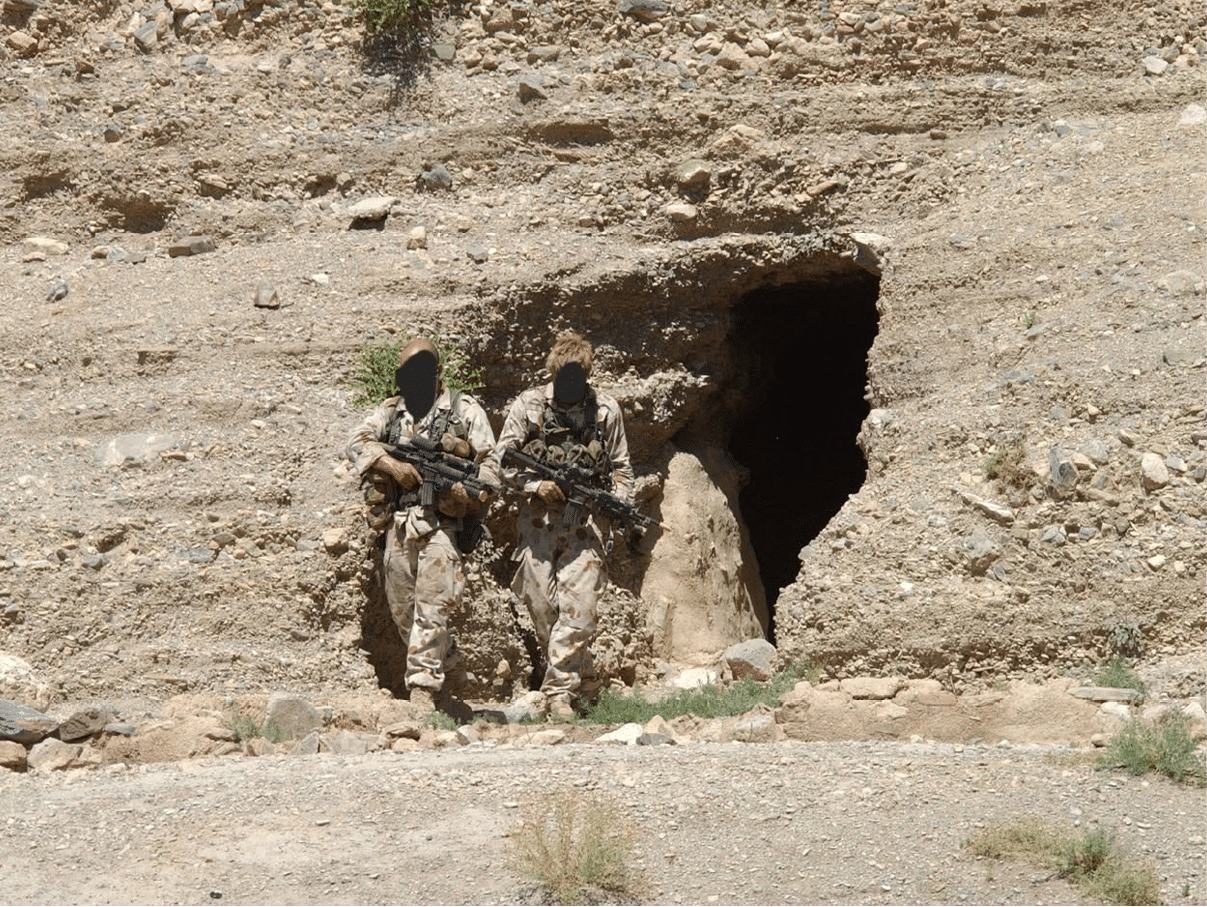 Two adults in military camouflage uniforms holding rifles stand near the entrance of a cave in a rocky, desert-like environment. Description generated by AI