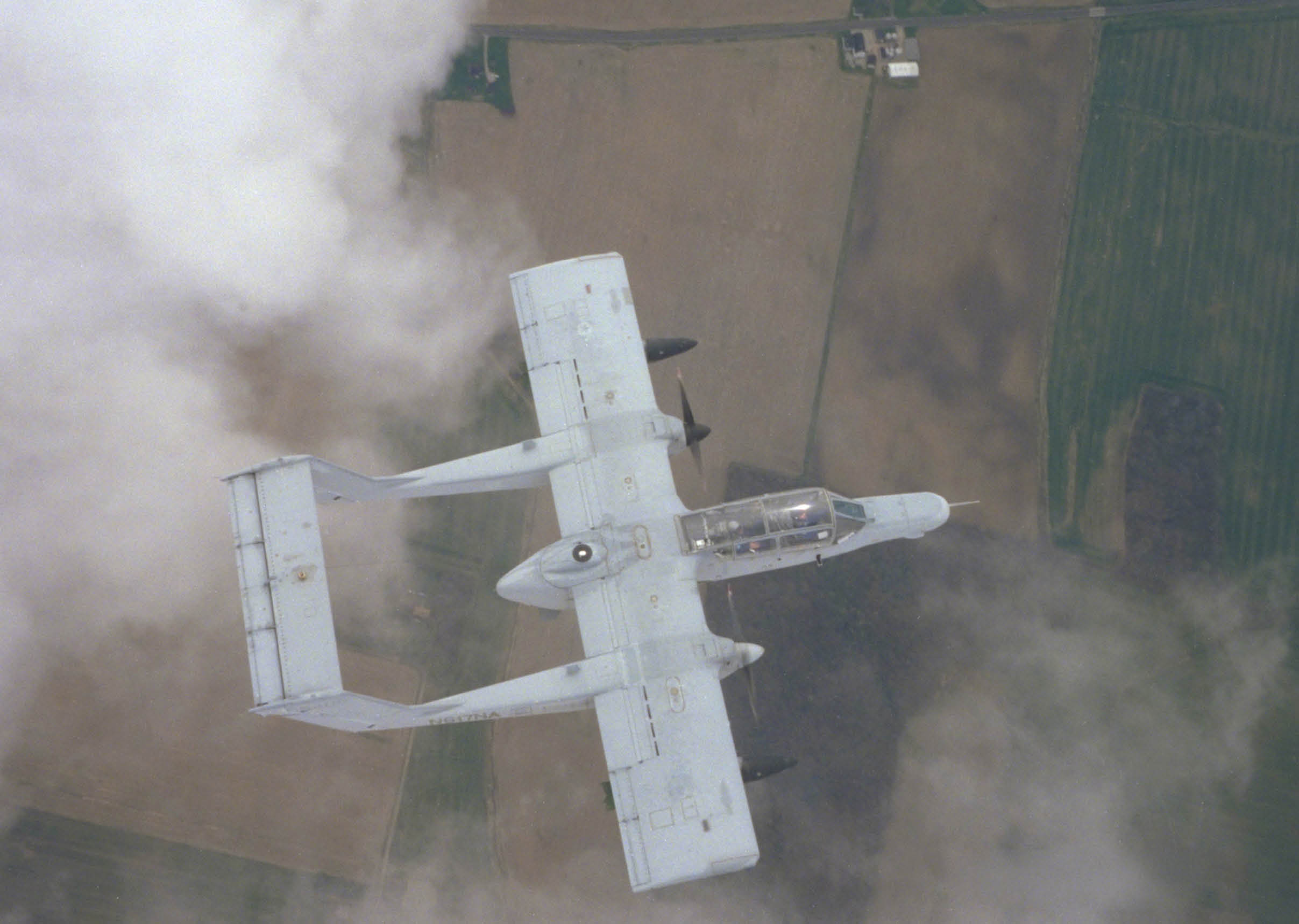 A military aircraft with twin booms and a central fuselage is flying above farmland and clouds. Description generated by AI