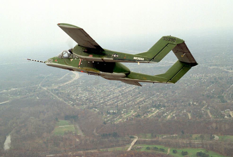 A green military aircraft with the tail number 55390 is flying over a suburban area with houses and trees below. Description generated by AI