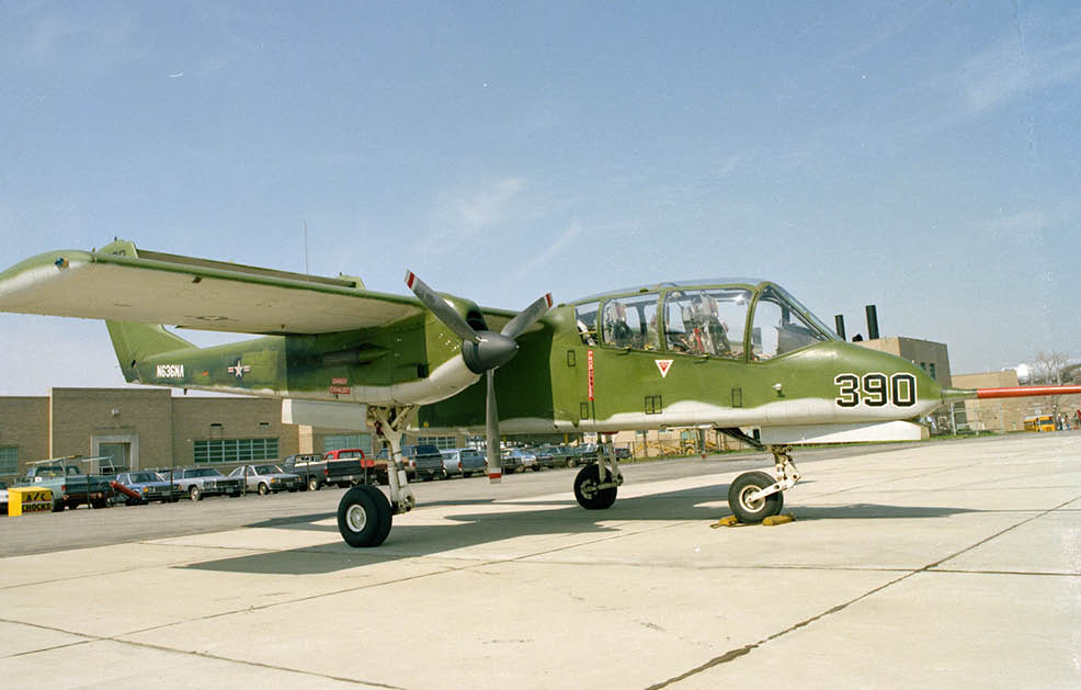 A green and white military aircraft with the number 390 on its nose is parked on a concrete surface near a row of cars and buildings under a clear sky. Description generated by AI