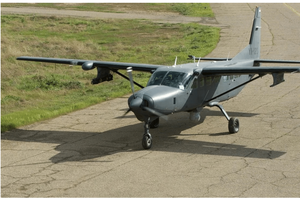 A small gray propeller airplane is parked on a cracked runway with grassy areas in the background. Description generated by AI