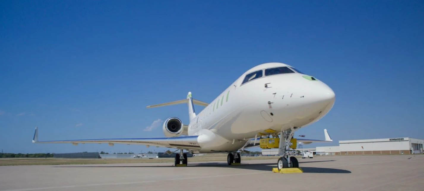 A white private jet is parked on a concrete runway under a clear blue sky with yellow wheel chocks placed around its front landing gear. Description generated by AI