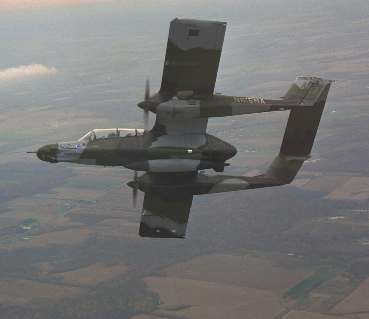 A military-style twin-boom propeller aircraft with camouflage paint flies over a rural landscape with fields and scattered clouds. Description generated by AI