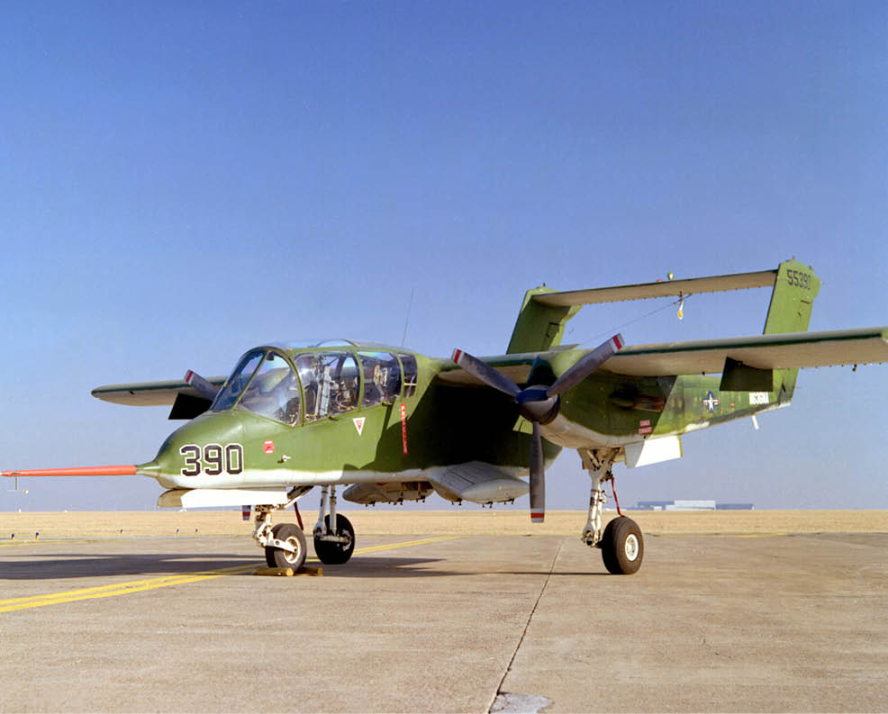 A green military aircraft with twin propellers is parked on a concrete runway under a clear blue sky. Description generated by AI