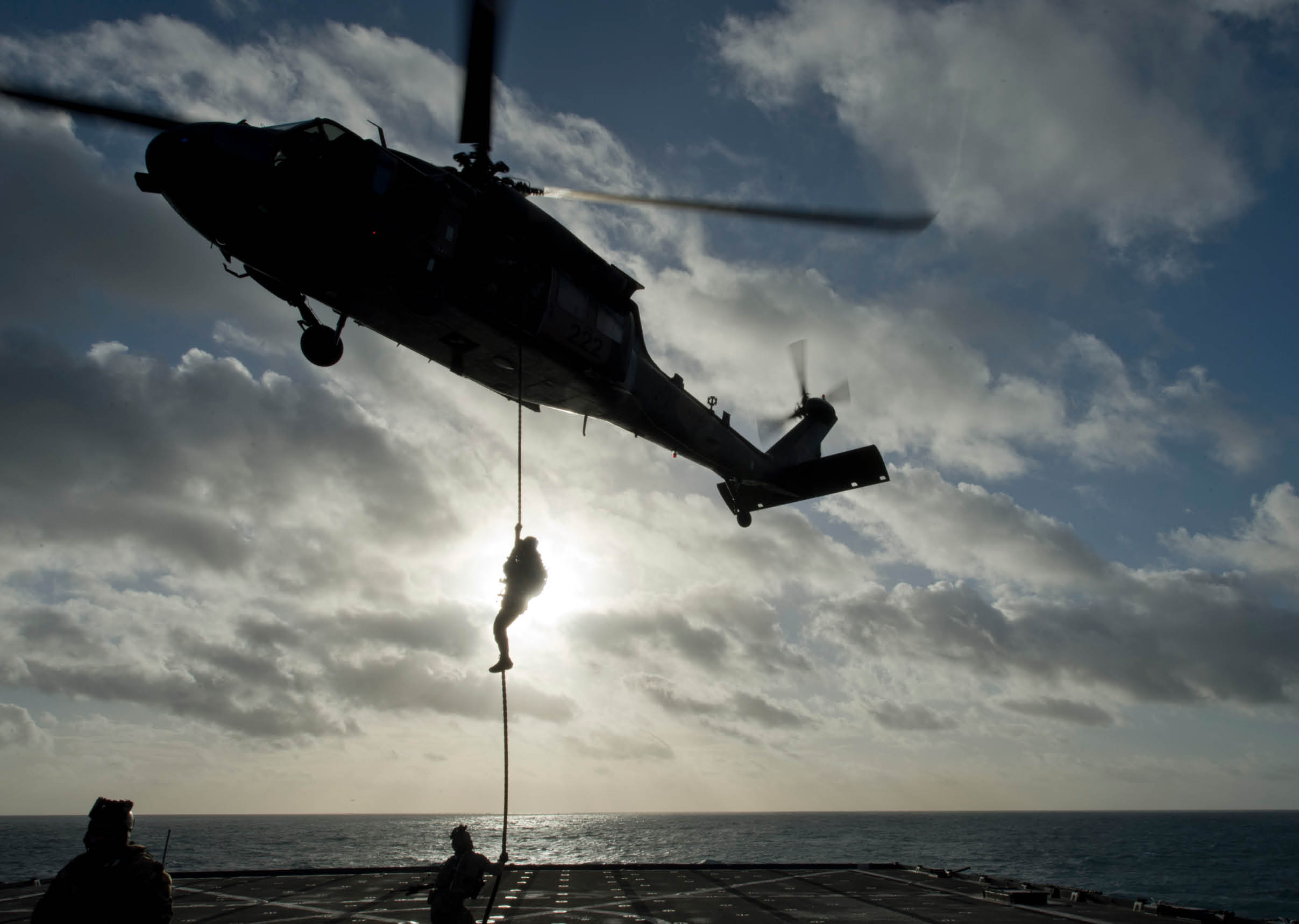 A helicopter hovers above a ship deck with one person descending on a rope while another person waits below. Description generated by AI