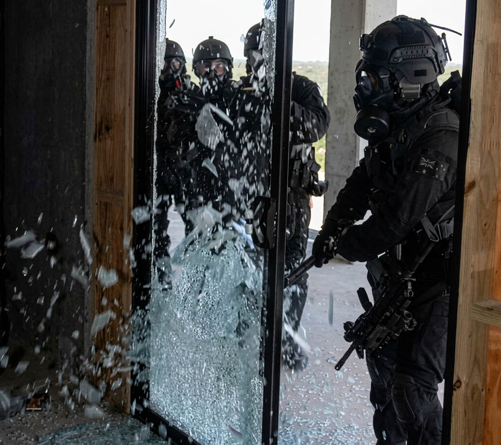 People in tactical gear are breaking a glass door with a baton in an indoor setting. Description generated by AI