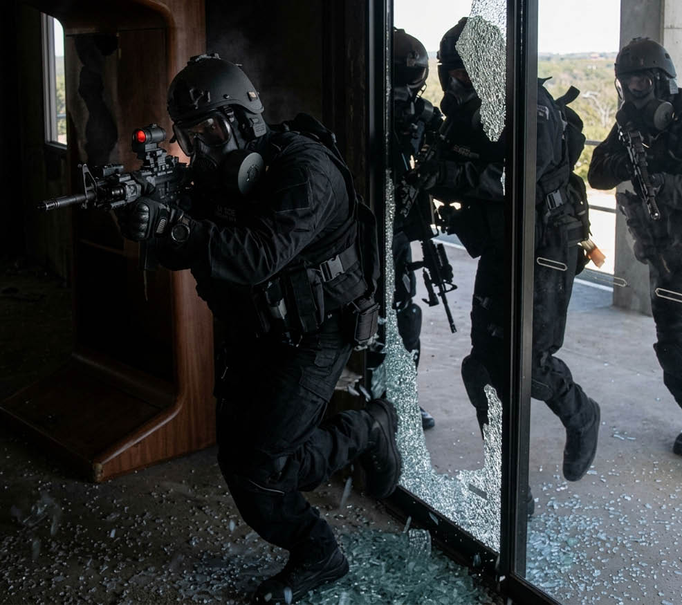 People dressed in tactical gear are moving through a broken glass door with weapons ready in a dimly lit indoor setting. Description generated by AI