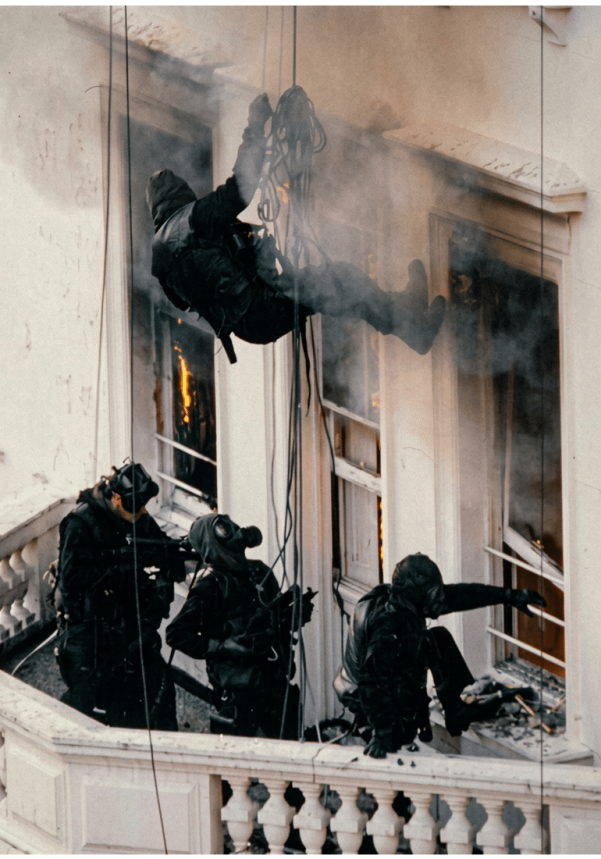 People in tactical gear and gas masks are rappelling and standing on a balcony of a building with visible flames inside. Description generated by AI