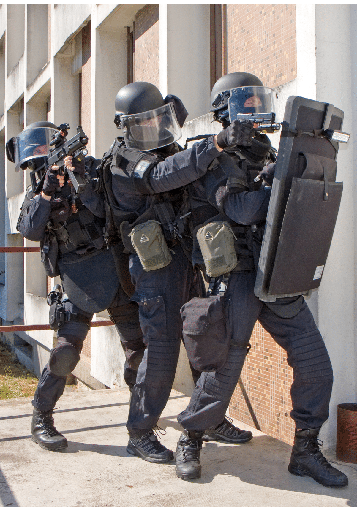 Three people in tactical gear and helmets are positioned closely together outside a building, aiming firearms with one holding a protective shield. Description generated by AI