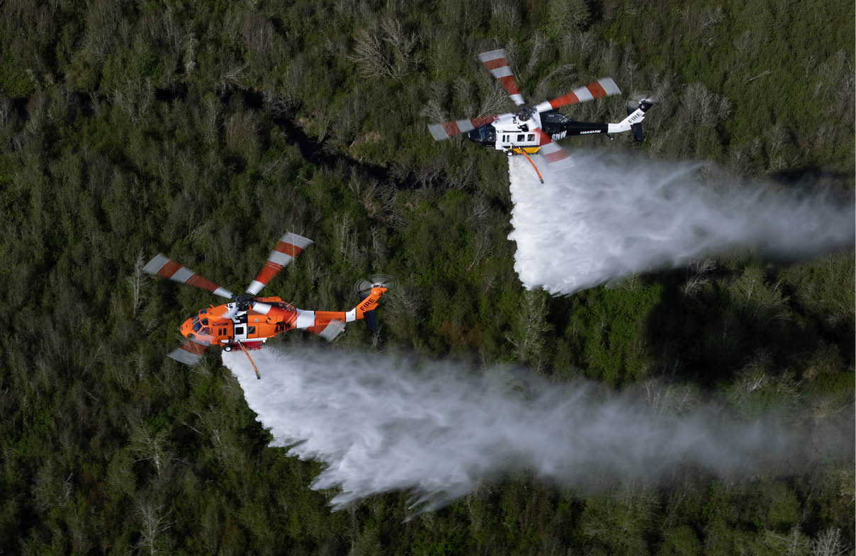 Two helicopters are flying over a forest while releasing large streams of water, likely to extinguish a wildfire. Description generated by AI