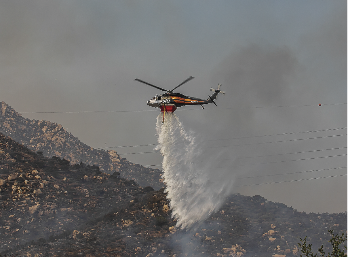 A helicopter is releasing water over a mountainous area, likely to extinguish a fire. Description generated by AI