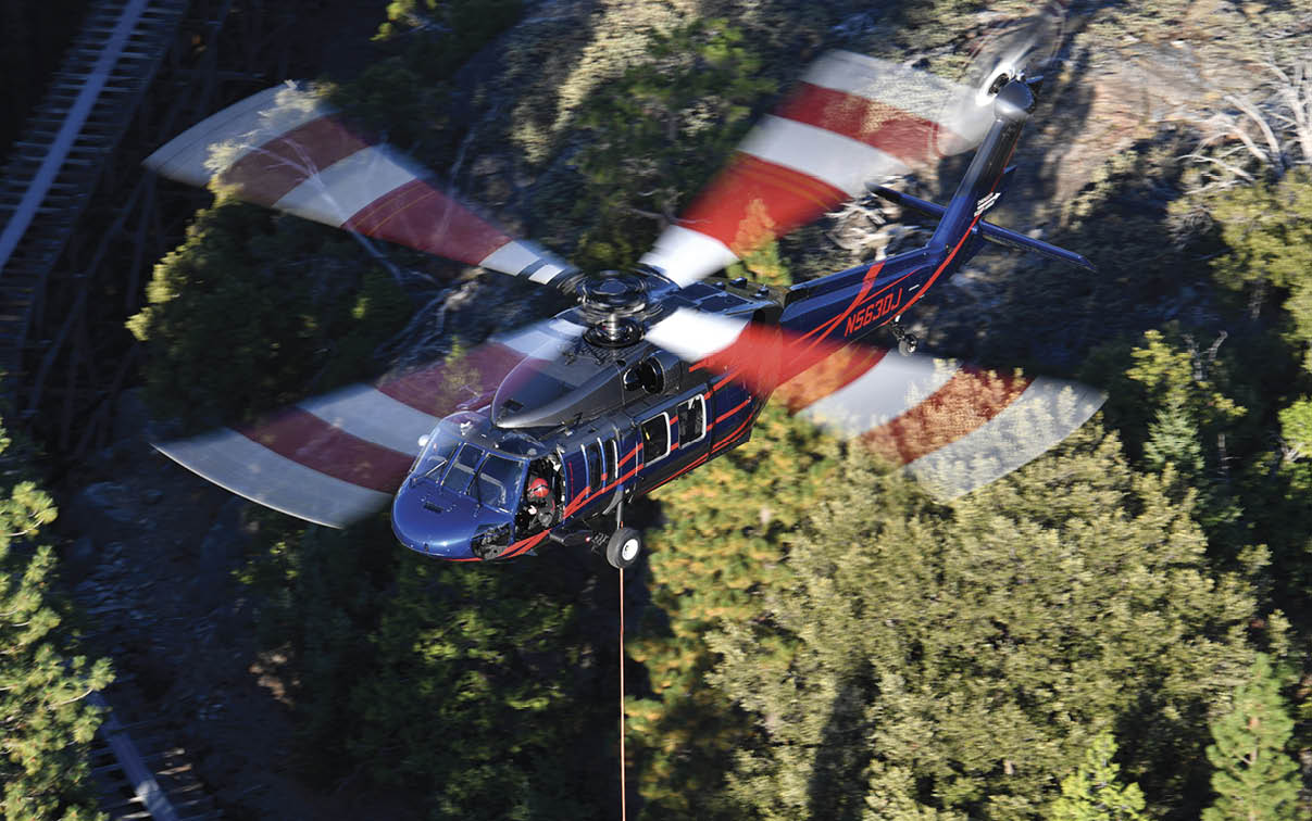 A helicopter with spinning rotor blades is flying above a forested area with a cable hanging below it. Description generated by AI