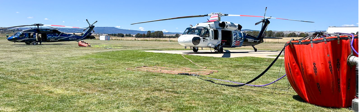 Two helicopters are parked on a grassy field. One helicopter is red and white, and the other is black and red. The red and white helicopter has a hose connected to it, possibly for refueling or maintenance. AI generated content